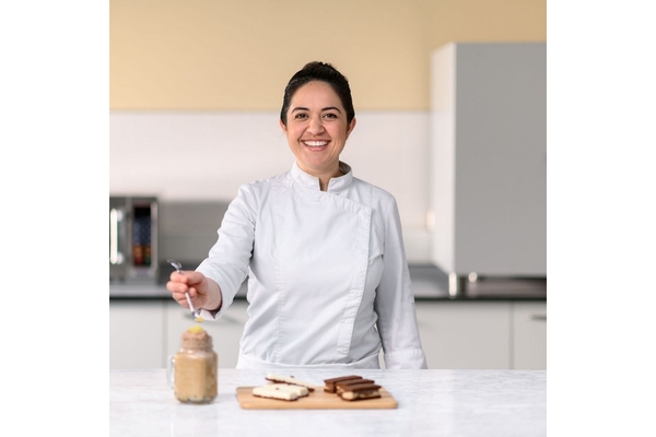 A chef in white apron smiling with cocoa powder and chocolates kept on kitchen countertop.